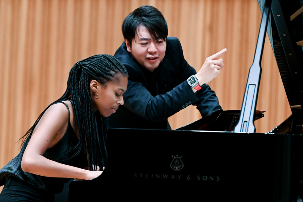 A black student, wearing smart attire, performing on the piano, with Lang Lang moving his arms behind her as he is teaching her, with a wooden panel behind them.